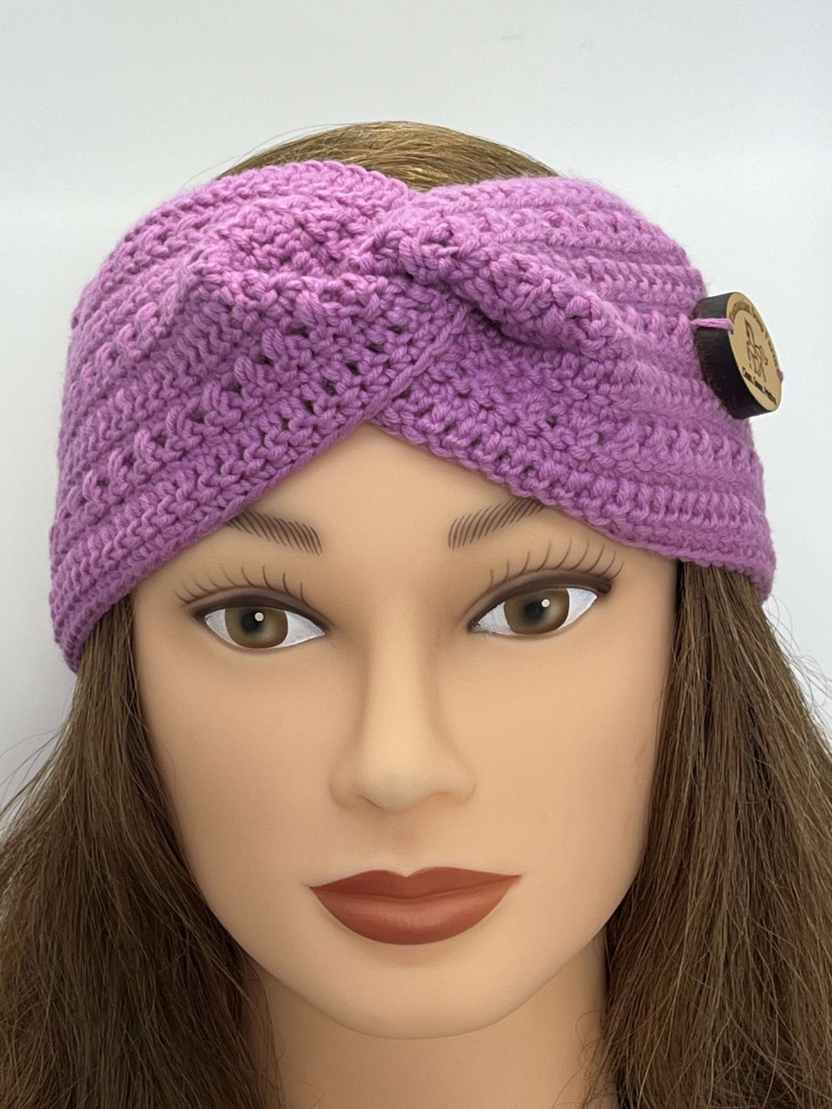 A pink crocheted headband by Tasmanian Soap Farm with a twisted knot design and a decorative wooden button, displayed on a mannequin head with brown hair against a plain white background.