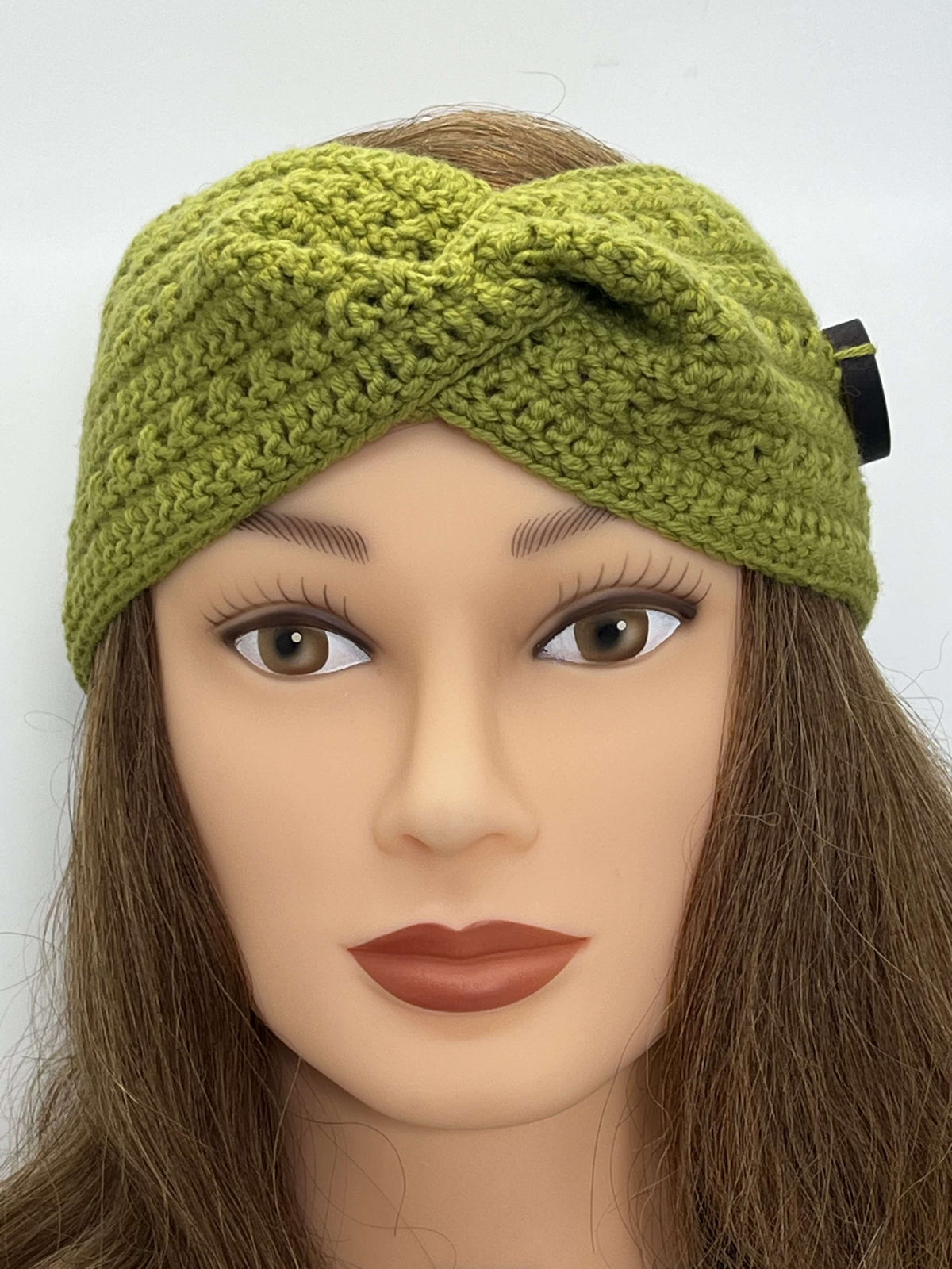 A grass green crocheted headband with a twisted front displayed on a mannequin head with brown hair against a plain white background.