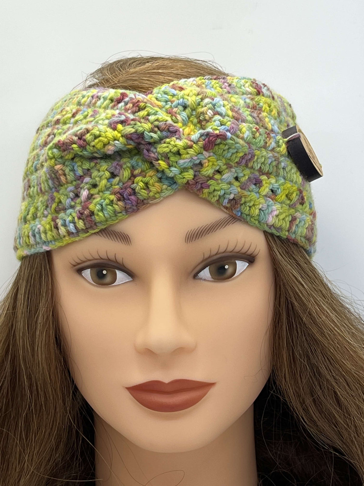 A multicolor crocheted headband with a twisted front detail and a small gold-tone button, displayed on a mannequin head with brown hair against a plain white background.