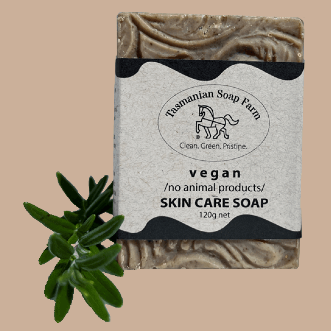 A rectangular bar of Tasmanian Soap Farm vegan skin care soap with a textured grey and brown marbled pattern, wrapped in a paper label. A small green leafy sprig is placed next to it on a solid beige background.