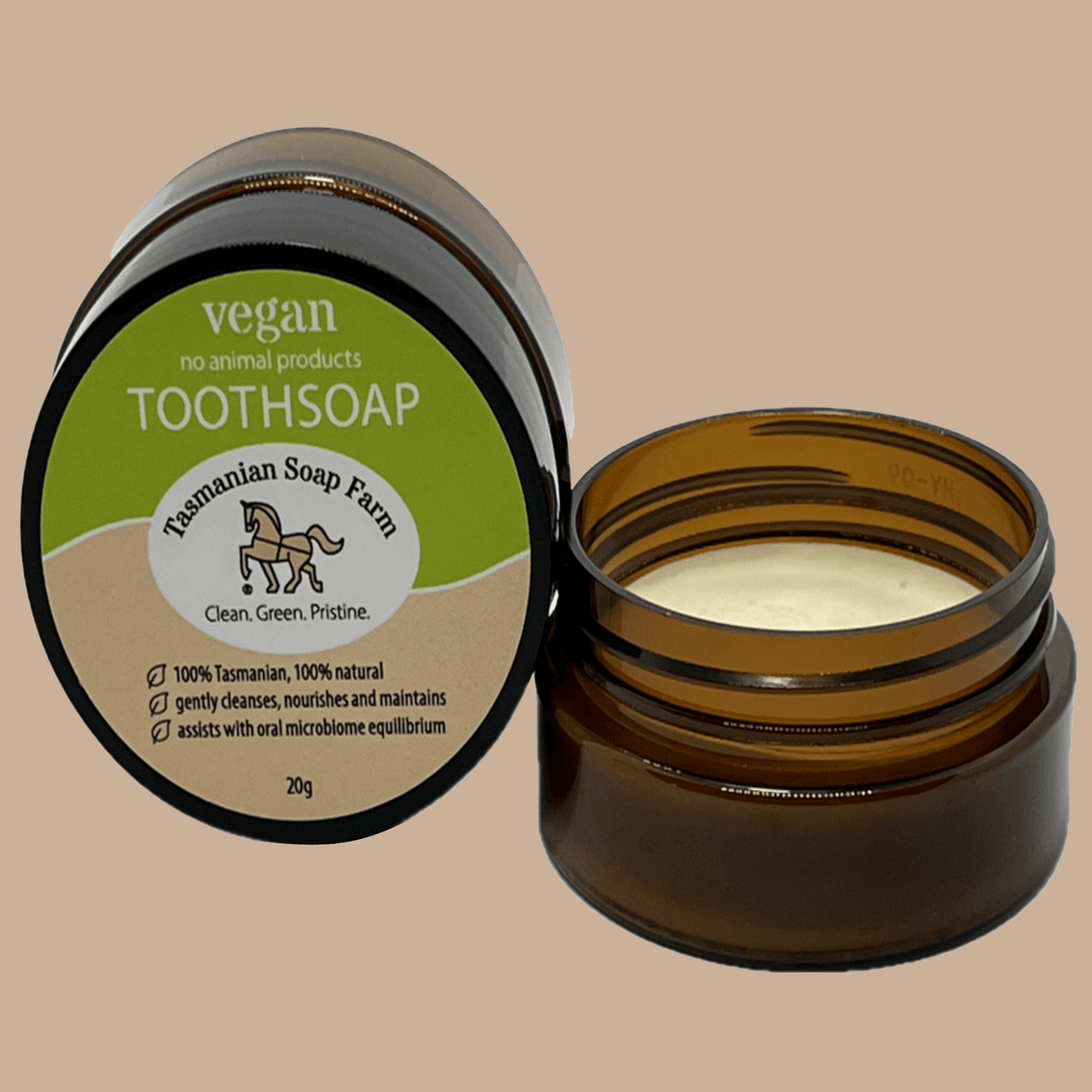 An open amber PET pot of vegan toothsoap next to its lid, which features a green and beige label for Tasmanian Soap Farm, set against a solid beige background.