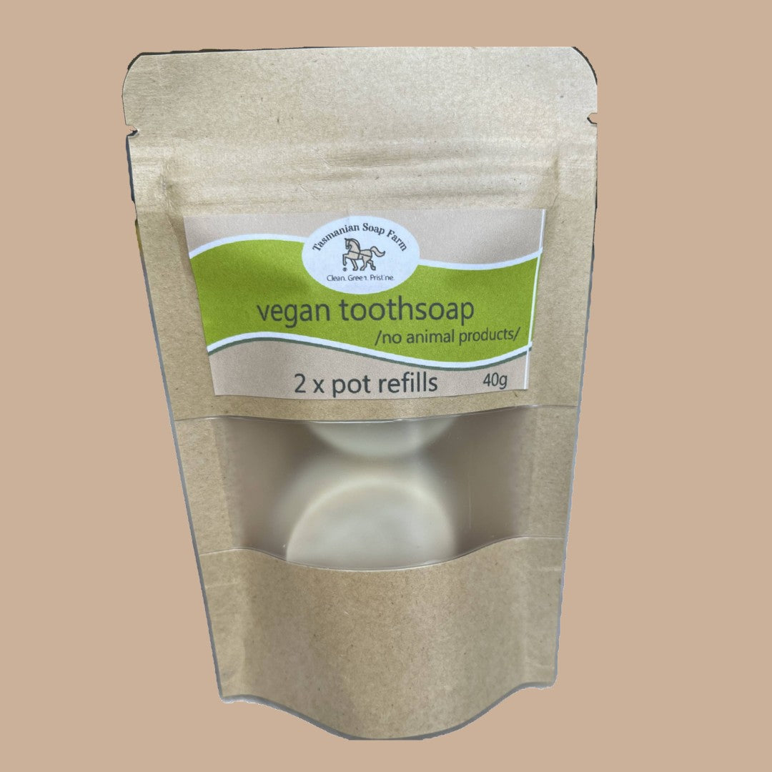 A kraft paper stand-up pouch of vegan toothsoap refill packs, featuring a green and white label with the brand 'Tasmanian Soap Farm' and a clear window showing two white bar soaps inside, set against a solid tan background.