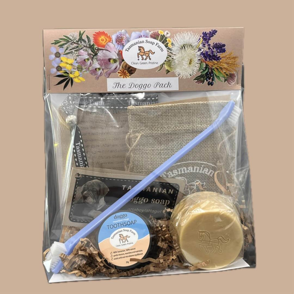 A packaged dog grooming kit labelled 'The Doggo Pack' by Tasmanian Soap Farm, containing a burlap pouch, a blue toothbrush, a round bar of tooth soap, and a round soap bar on a white card background.