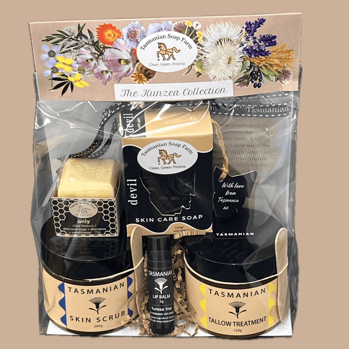 Tasmanian Soap Farm gift pack, 'The Kunzea Collection' set in clear cello packaging, featuring a bar of soap, two  jars (skin scrub and tallow treatment), a black tube of lip balm, shampoo soap and a soap net, all arranged on a buff background.