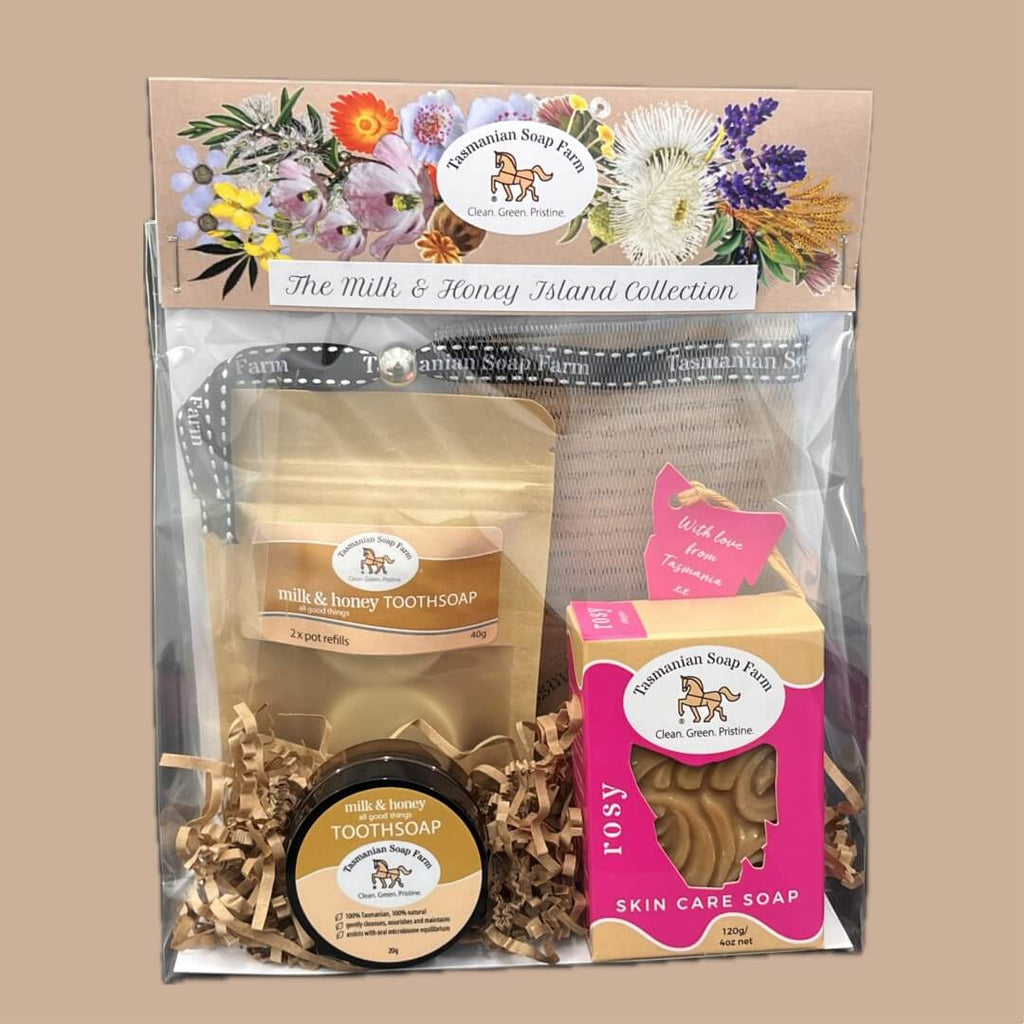 A packaged gift set, The Milk & Honey Island Collection, by Tasmanian Soap  Farm, containing milk and honey toothsoap, a small jar of toothsoap, a pink box of Rosy skin care soap, and a mesh bag, all arranged in a clear bag with shredded paper and a floral header card.
