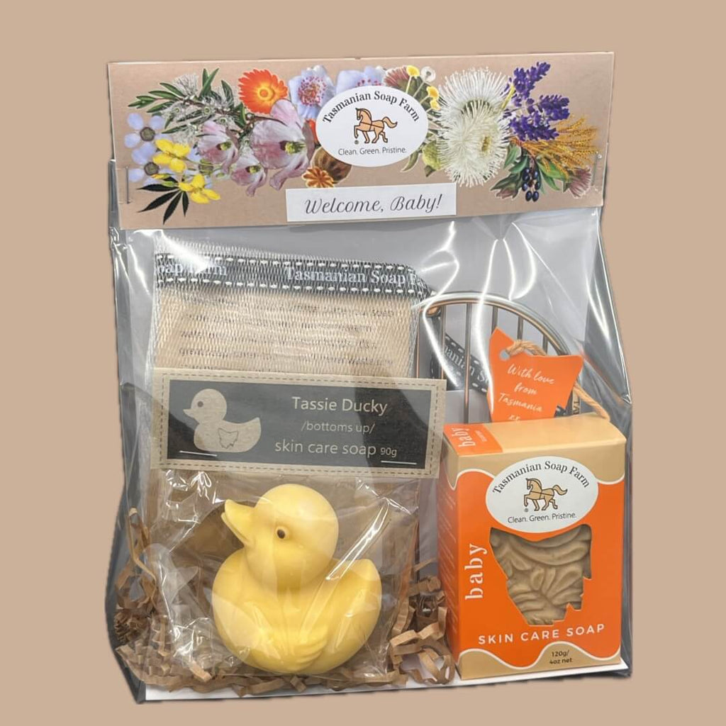 A baby-themed Tasmanian Soap Farm gift set in a clear cello bag, featuring a yellow duck soap, a baroque-textured baby skin care soap bar in an orange box a stainless steel soap rack and a soap net, all under a floral header card.
